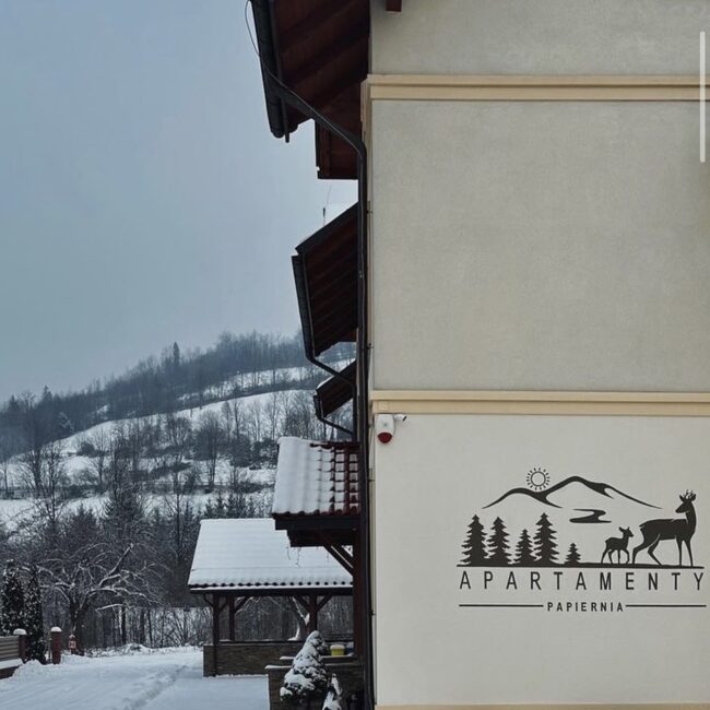 Budynek Agroturystyczny Papiernia w zimowej scenerii, z logo i grafiką na elewacji, idealne miejsce na spokojny wypoczynek w górach zimą.
